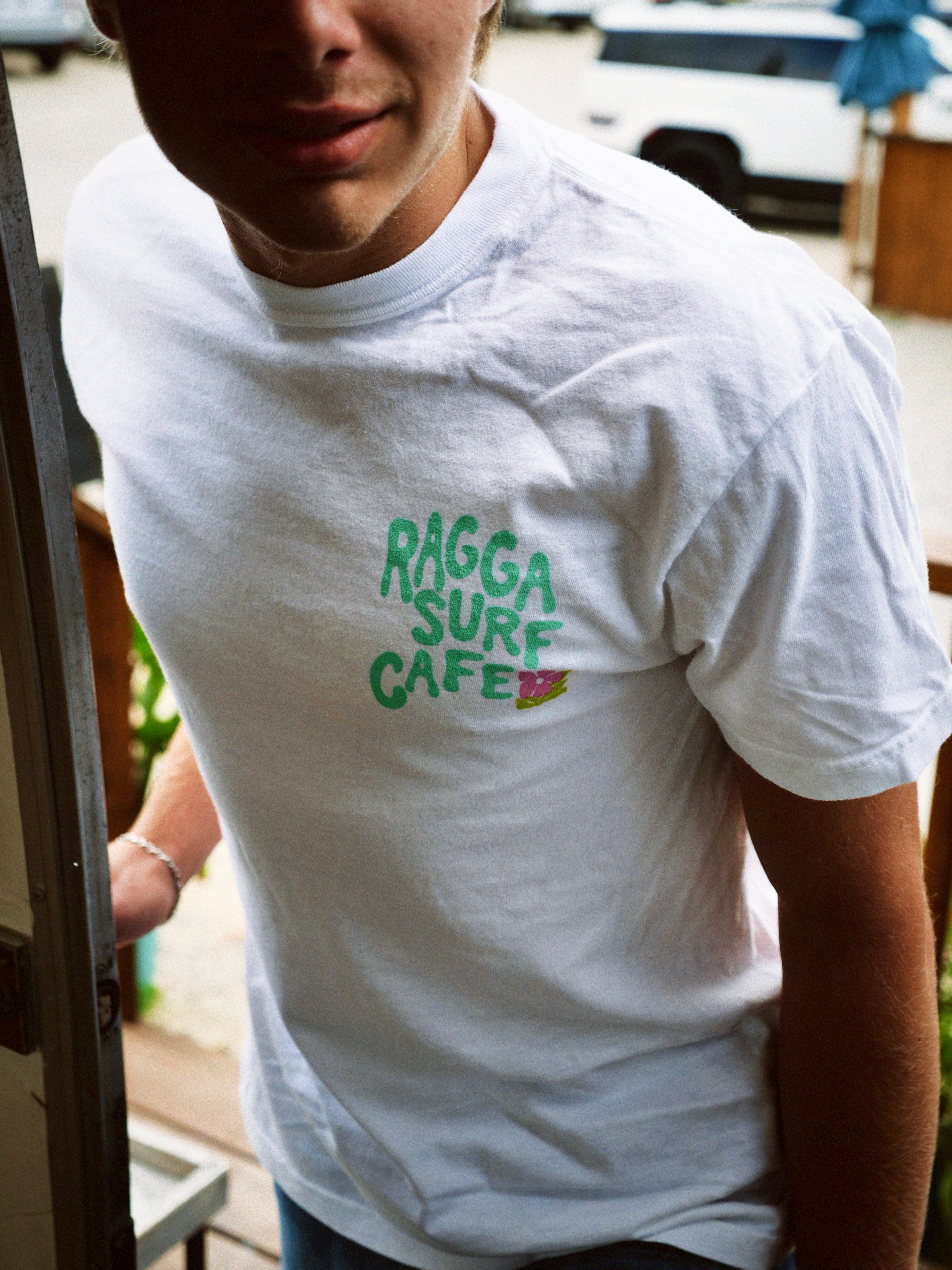 Person wearing a white t-shirt with 'Ragga Surf Cafe' text in green.