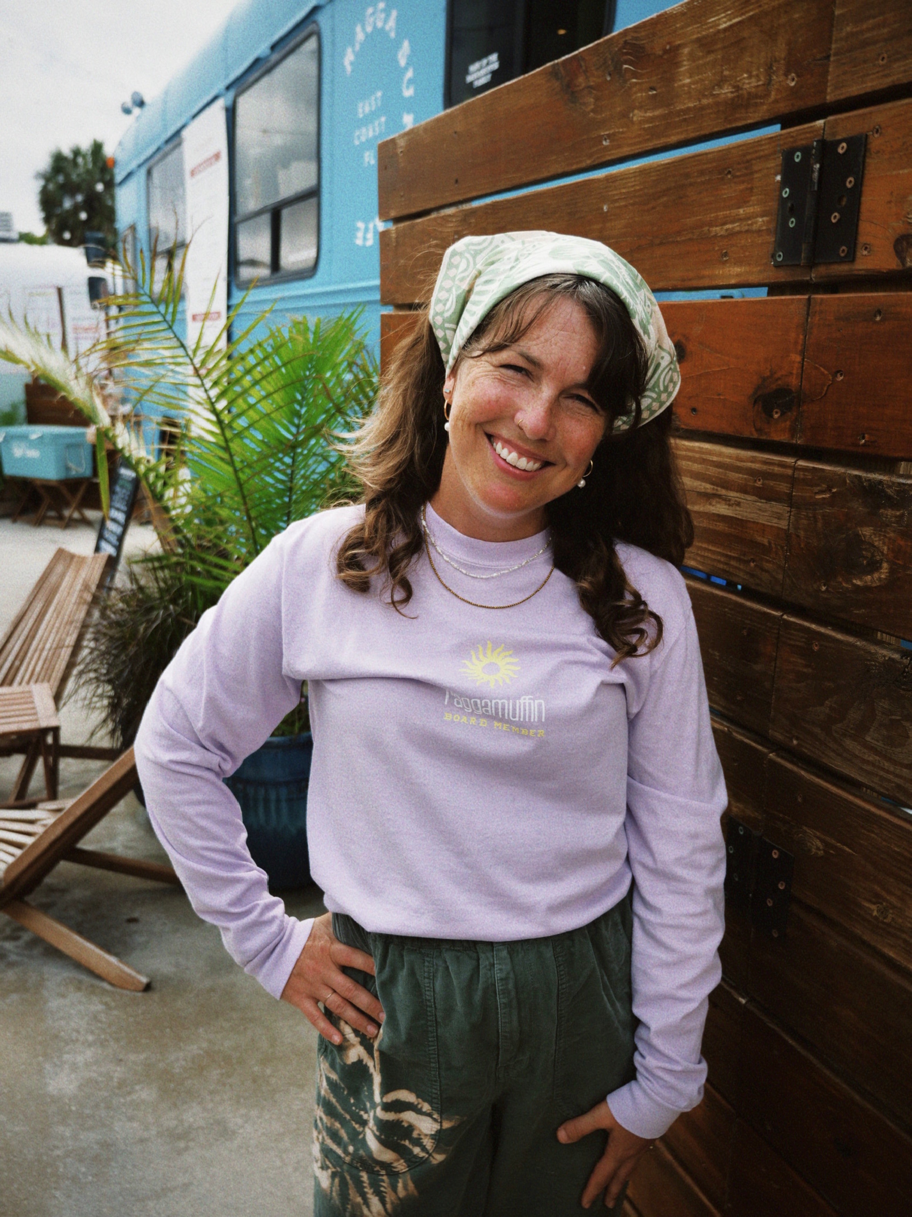Woman wearing a light purple long-sleeve shirt with a logo, standing outdoors near a wooden bench and a blue food truck.